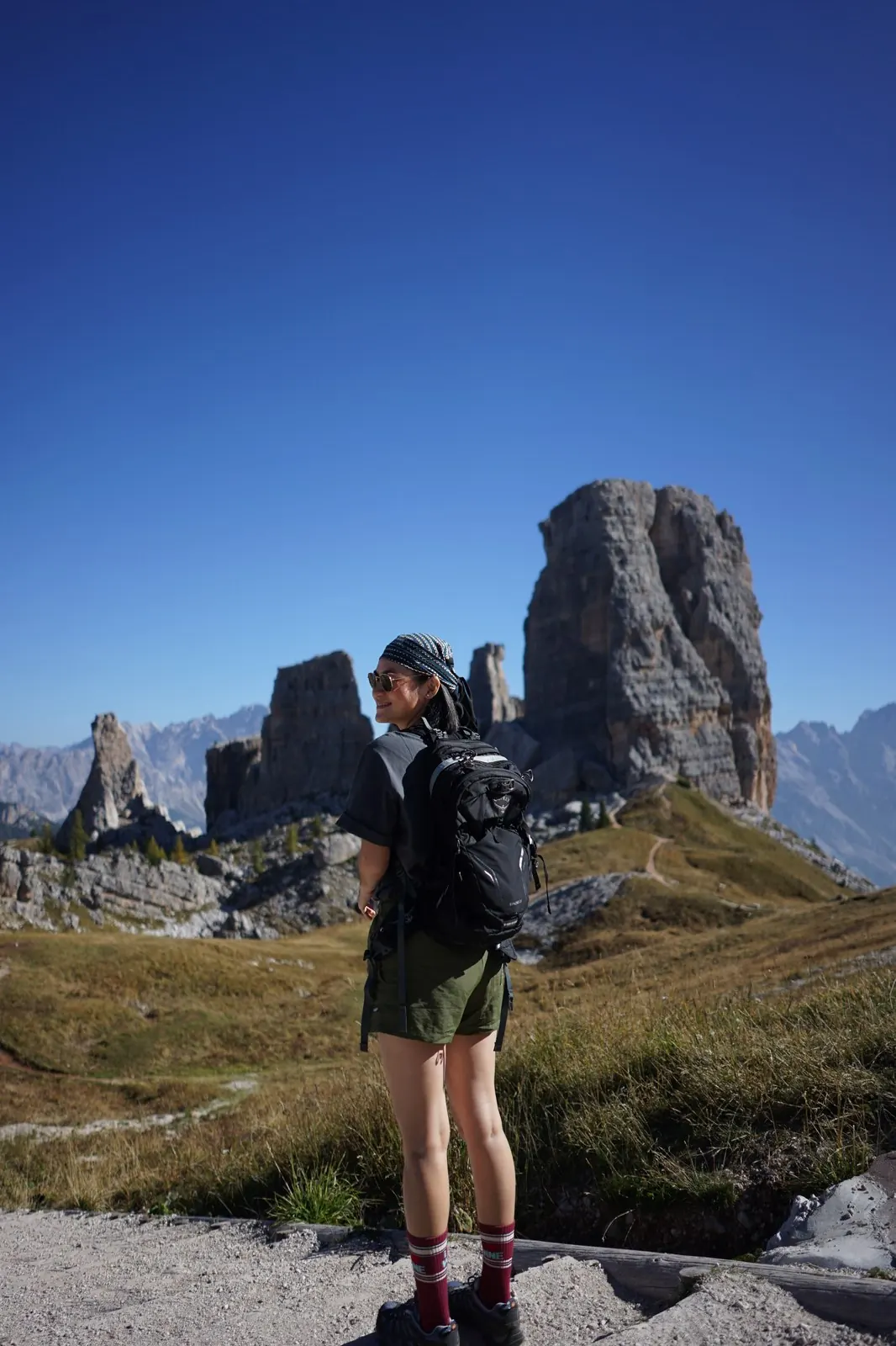 Milk with her backpack smiling on a trail in the Dolomites, Italy, towering rock peaks behind her