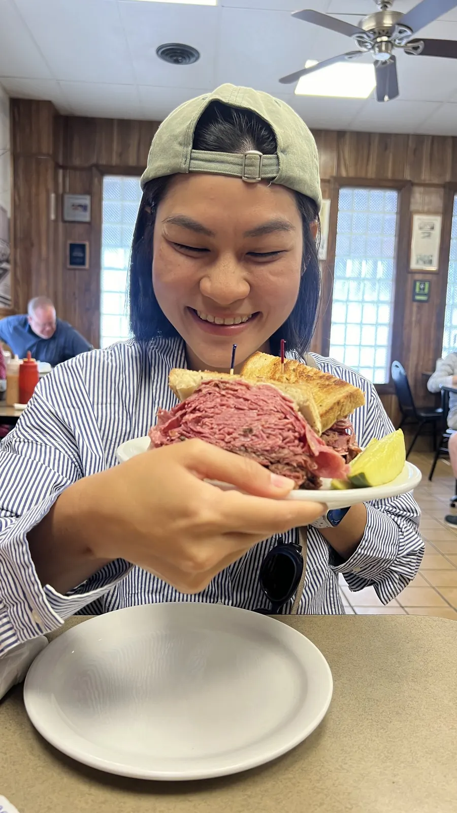 Milk in a striped shirt and backwards cap, beaming as she holds up a plate with a giant deli pastrami sandwich
