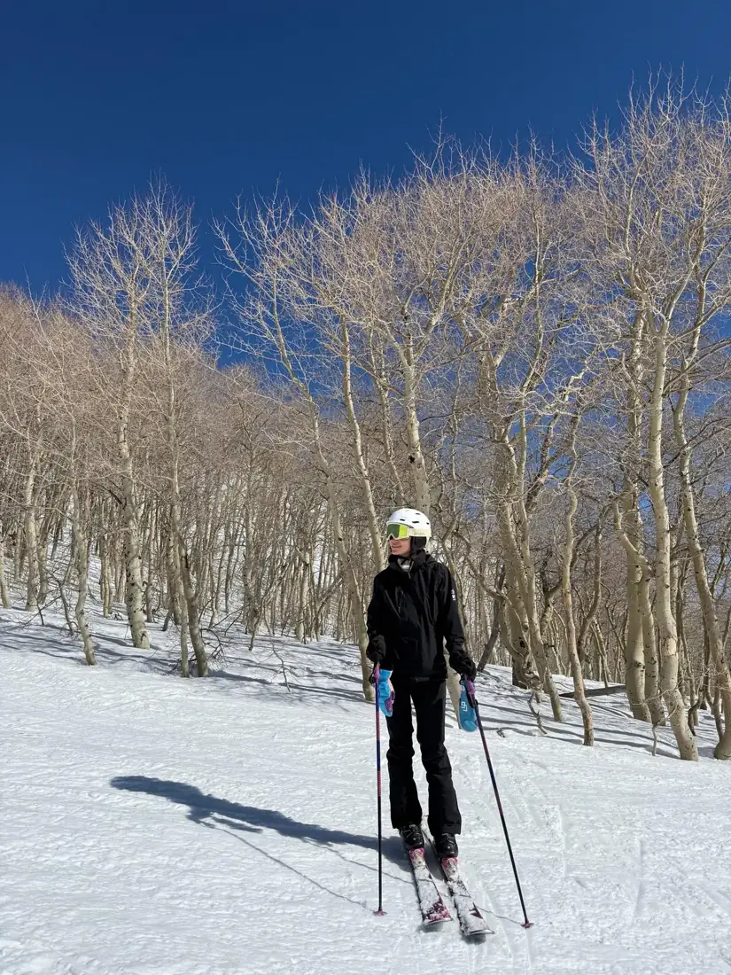 Milk in black ski gear and a white helmet, green goggles pushed up, standing on her skis among bare aspen trees under a bright blue sky in Utah