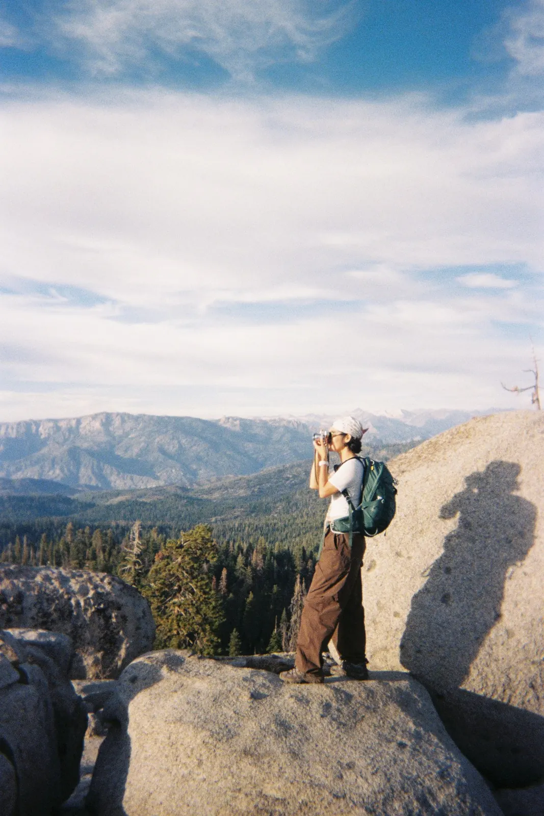 Milk on a mountain summit in Yosemite, backpack on, holding her film camera to her eye with peaks and forest behind her