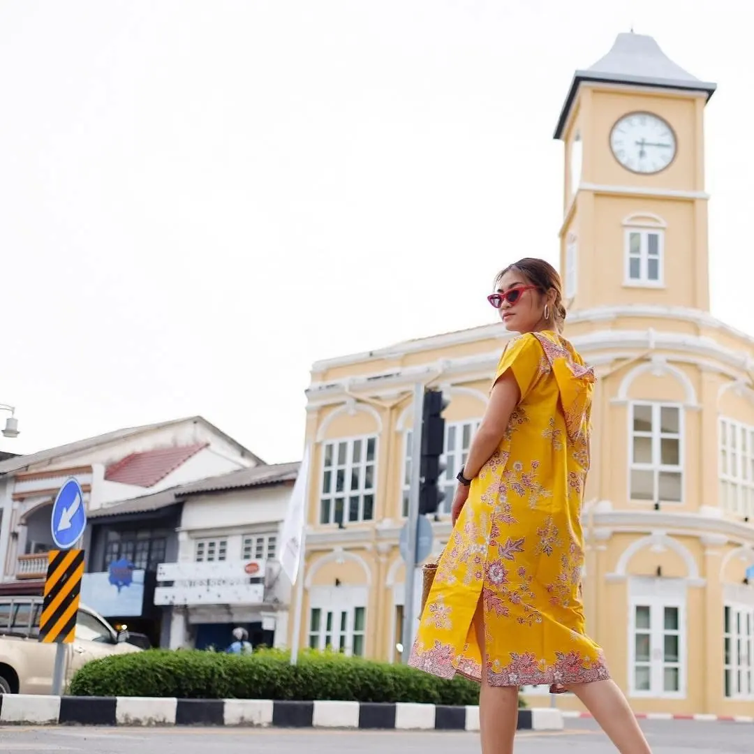 Milk wearing a yellow Phathoongnoongdai batik dress with pink florals, walking past the clock tower in Phuket Old Town